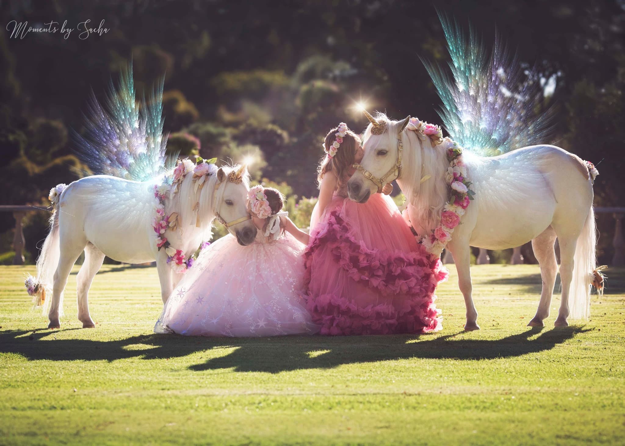 Two girls in pink gowns with two flower-adorned unicorn ponies on the manor lawn